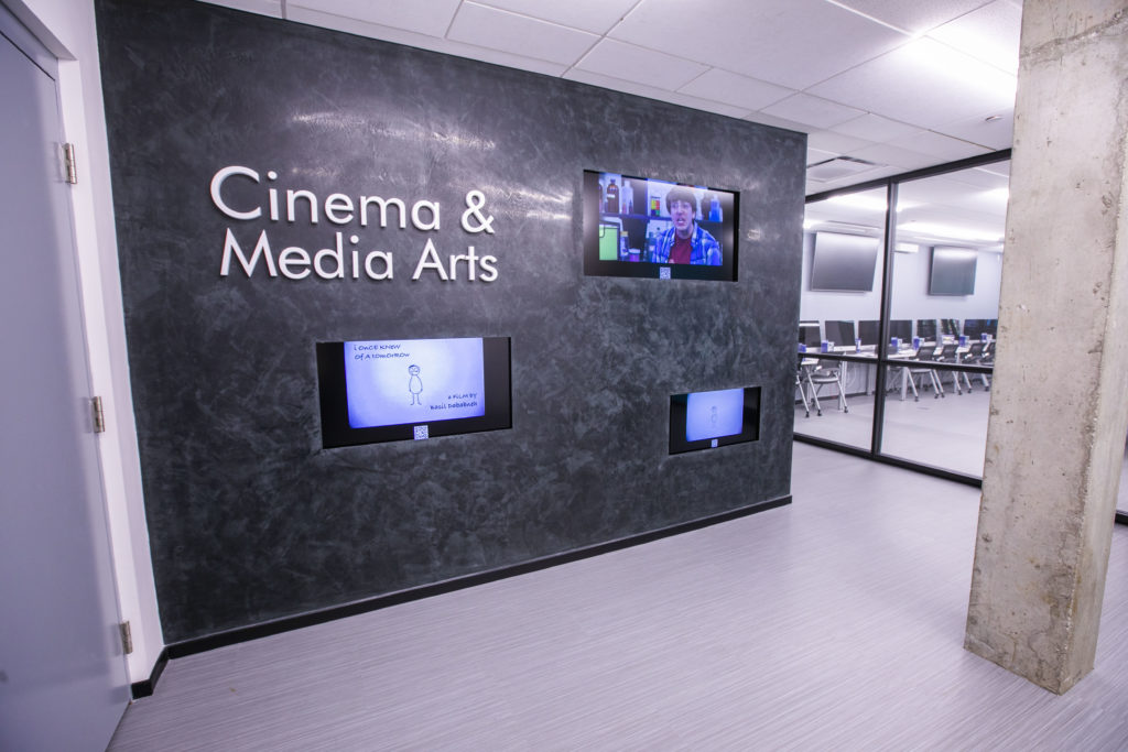 The wall of screens outside the Buttrick elevator in the basment with a sign saying "Cinema & Media Arts" and three TV screens showing student films