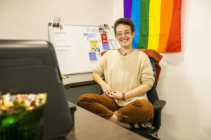 Cassidy Parkison, a white woman with short brown hair wearing a beige sweater and sitting cross-legged in an office chair with a pride flag behind her