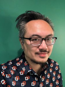 A headshot of Iggy Cortez, an Asian man with glasses and dark pushed back hair and short facial hair standing against a green background wearing a patterned shirt
