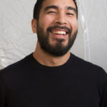 A headshot of Alejandro Acierto, a brown-skinned man with dark hair and beard who is laughing and wearing a black shirt
