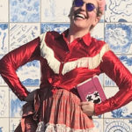 A headshot of Krista Knight, a white woman with pink hair wearing sunglasses and a red western shirt standing against a blue tile wall with her hands on her hips