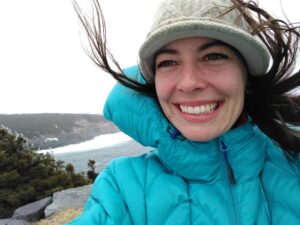 Tamara Carley, wearing a blue puffer jacket and khaki hat, standing on a windy beach with the ocean and hills in the background