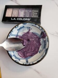 Closeup photo of a soy sauce dish with a grapefruit spoon in the foreground with a mixture of purple eyeshadow mixed into a paste with olive oil. An eyeshadow color palette is in the background with shimmery shades of pinks and purples.