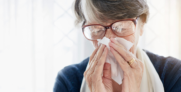 An old woman blows her nose, holding the tissue tightly, her eyes tight shut. She may be sneezing or weeping.