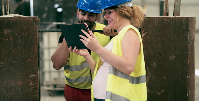 Pregnant woman and male coworker wearing hard hats and safety vests looking at ipad in factory