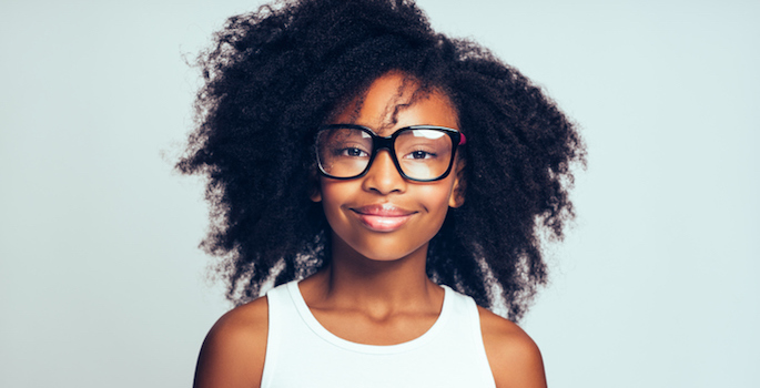 Smiling young African girl with long curly hair wearing glasses while standing alone against a gray background