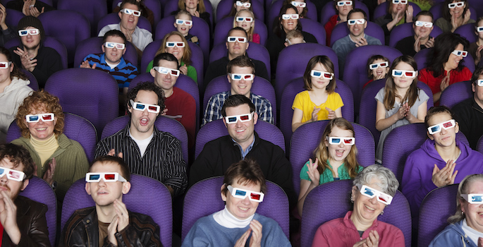 A large audience wearing 3D glasses in a darkened movie theater.