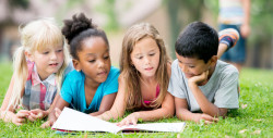A multi-ethnic group of elementary aged children are reading a book in the park.