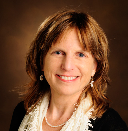 woman with light brown hair, wearing white blouse, necklace, and black sweater