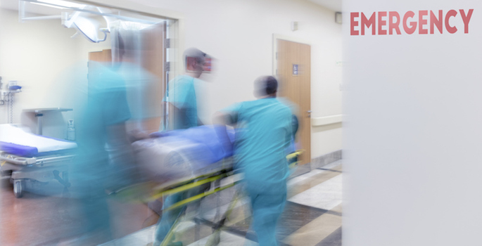 Three emergency medical workers rushing patient in gurney down hall past sign reading 