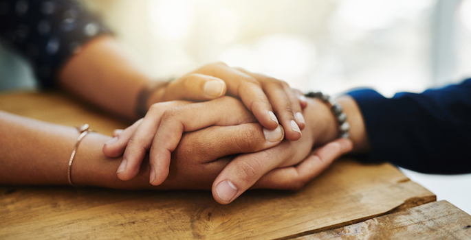 Close up of a person's hand being clasped in a comforting manner between the hands of another person.