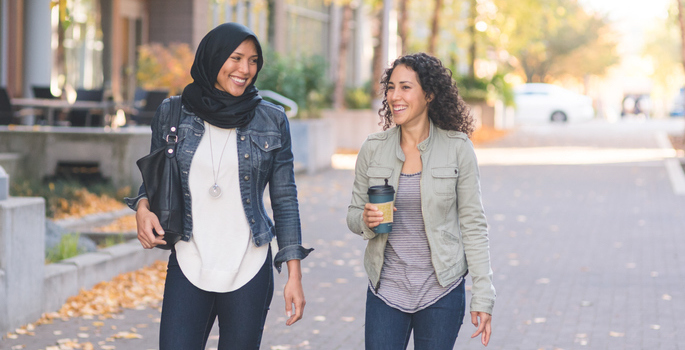 Two women of middle eastern descent, one wearing hijab, strolling down the sidewalk deep in conversation.