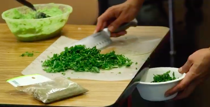 close-up of hispanic woman's hand sweeping chopped herbs off a cutting board into a bowl with the flat edge of a knife