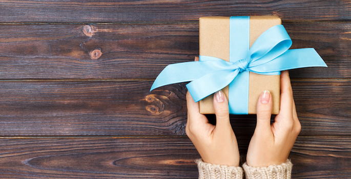 caucasian woman's hands holding a small gift wrapped in brown paper with a blue bow against a dark wood paneled background