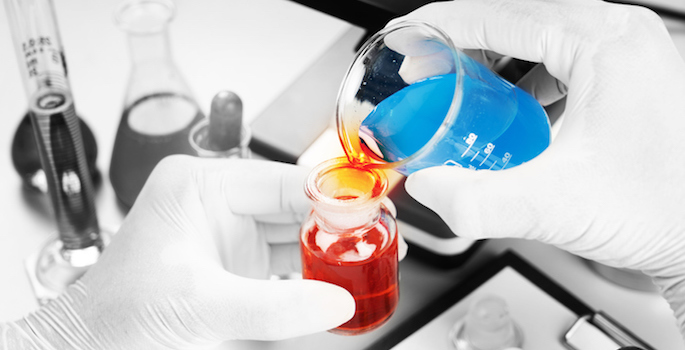 close up of gloved hands in lab pouring blue liquid into beaker containing red liquid