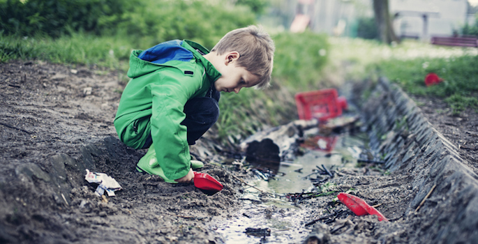 Little boy in green raincoat digging intently in mud puddle