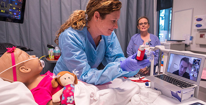 Nurse in protective gear leans over pediatric simulation dummy holding an inhaler medication up to a computer screen for a male nursing student, who is teleconferencing in, can see the label. The nursing instructor stands in the back of the room, observing.
