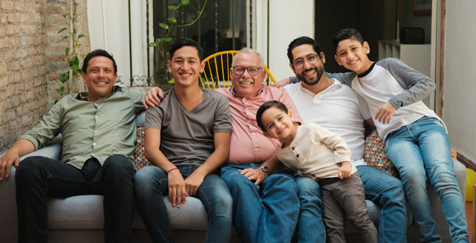 A Latino grandfather sitting on an outdoor sofa surrounded by his sons and grandsons