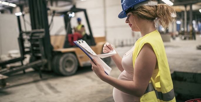 White heavily pregnant woman wearing a blue hard hat and yellow vest looks at a clipboard in a warehouse while a colleague drives a forklift behind her.