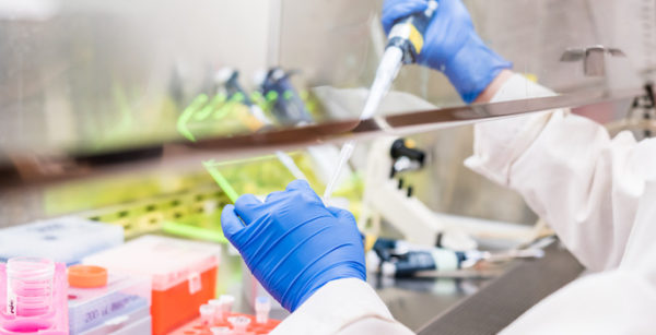 Close up photo of white-coated, gloved hands extracting fluid from a vial underneath a fume hood in a laboratory