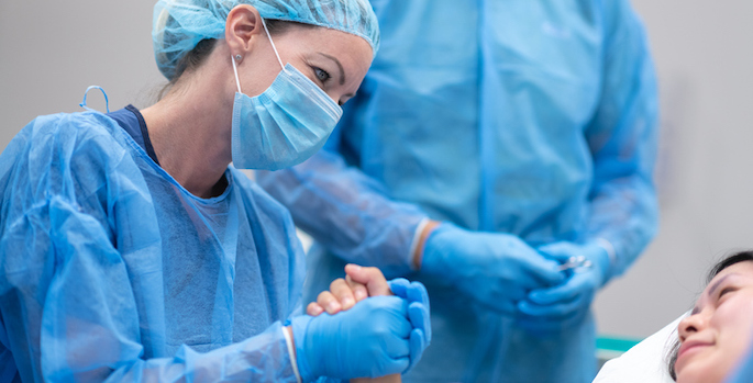 Woman in blue surgical wear, including hair net and mask and gloves (PPE), holding the hand of a patient in bed. Another healthcare provider wearing PPE stands behind them.