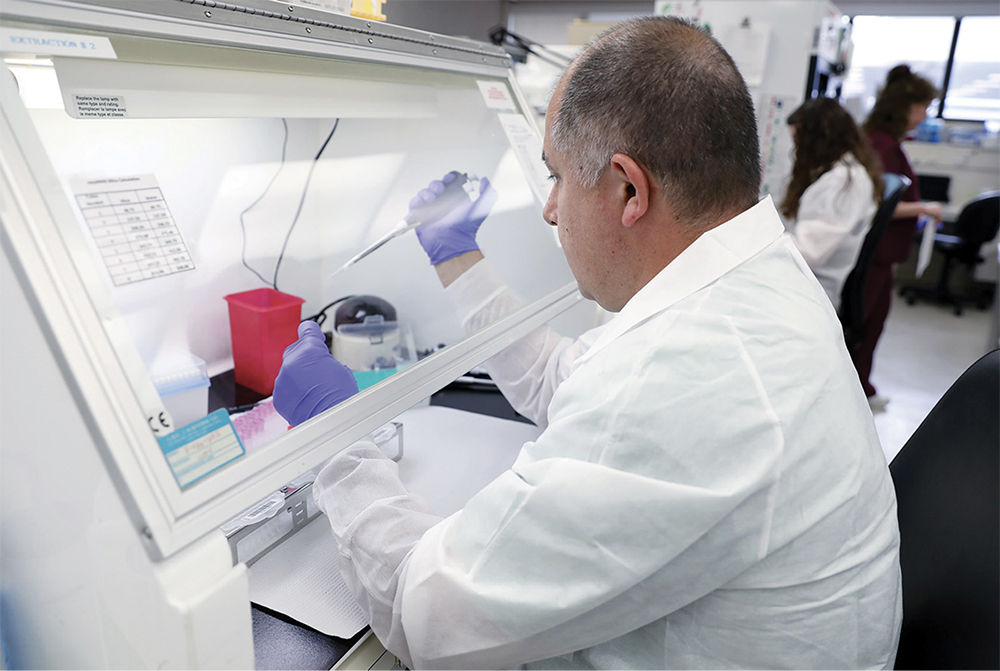 photo of Giovanni Suarez examining COVID-19 specimens from behind a safety screen