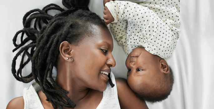 Black South African mother with artfully arranged dreadlocks and white tank top lying in bed with her newborn wearing a white onesie with black polka-dots. Mom and baby are facing each other and smiling.