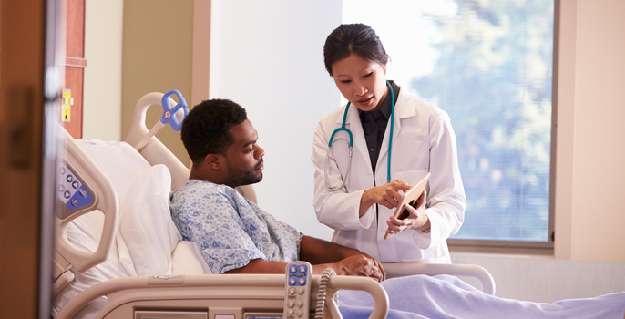 Asian American female doctor discusses care with hospitalized young African American male patient by showing him information on a digital tablet