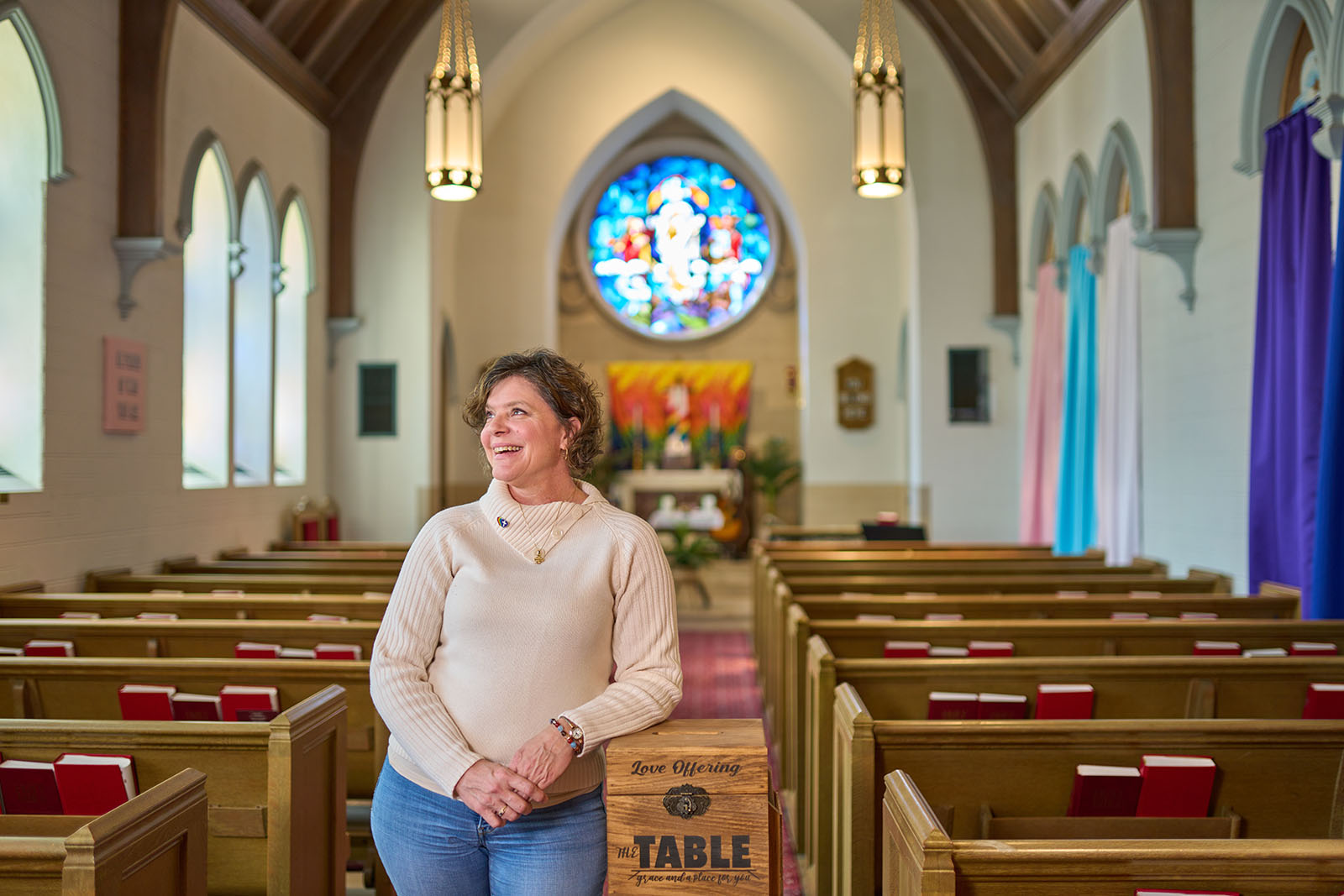 Pastor Dawn Bennett in white sweater and jeans in the middle aisle of her church in downtown Nashville with the altar and stained glass window behind her.
