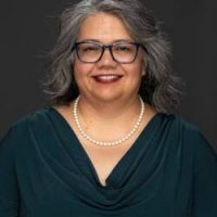 woman in dark blue blouse, pearls, glasses, with black and gray hair