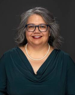 woman in dark blue blouse, pearls, glasses, with black and gray hair