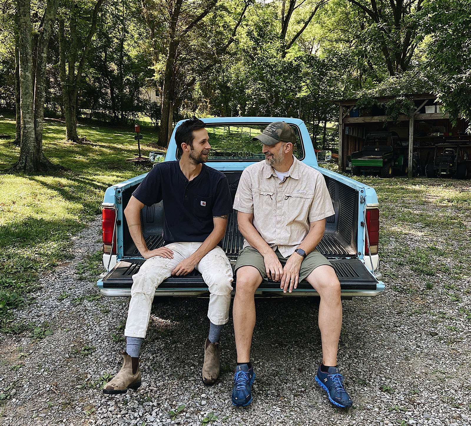 A young man and his father sitting on the back of a truck in a wooded setting.
