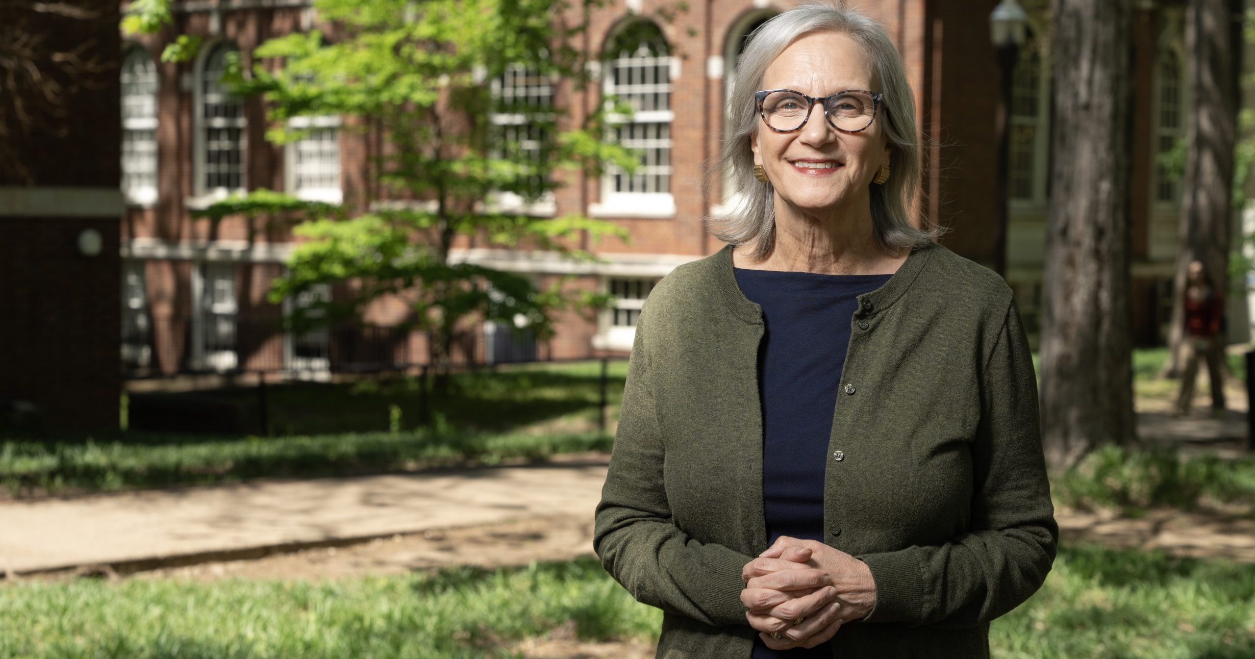 A woman in glasses standing in front of a brick building and trees.