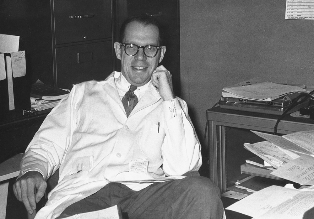 Black and white photo of a grinning Dr. Blair Batson in his messy office