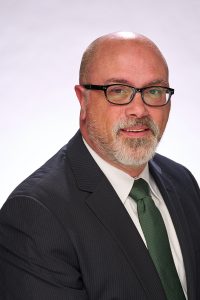 bearded bald man wearing black glasses black suit jacket white shirt and green tie