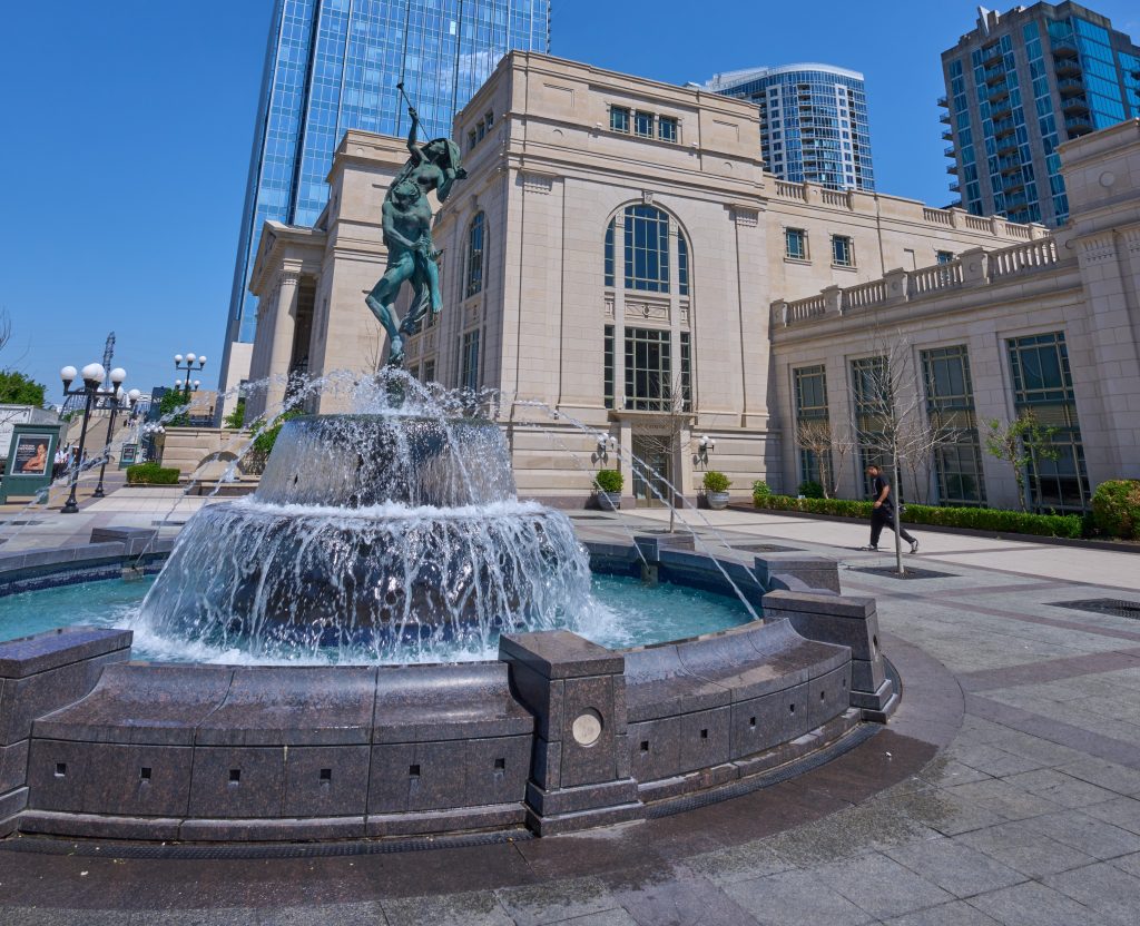Fountain with bronze statue in front of the Schermerhorn Symphony Center in downtown Nashville, with the Pinnacle building and other high-rises in the background on a clear blue day.