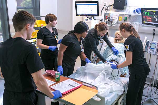 Nursing students in black scrubs around a mannequin in a simulated hospital bed setting