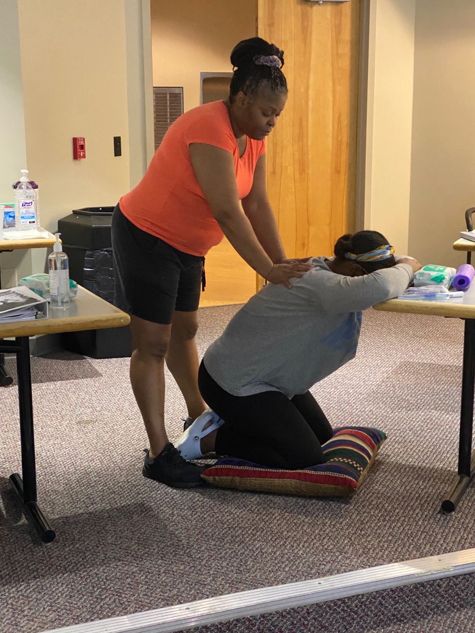 two people, one kneeling on a pillow and the other pushing on her shoulders