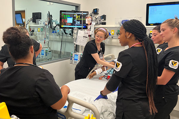 5 students in black VUSN scrubs circle a hospital bed in the sim lab