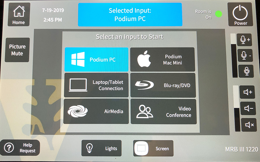 Photo of the control panel, which shows a menu of input options: Podium PC, Podium Mac Mini, Laptop/Tablet Connection, Blu-ray/DVD, AirMedia, and Video Conference. Additional options are located on the periphery of the screen.