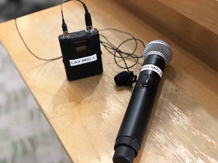 Photo of a handheld microphone and a clip-on microphone sitting on a wooden surface. The clip-on has a label that says "LAV MIC 1," a cord with a mic at the end, and an antena. The handheld mic is labeled "HH MIC 3."