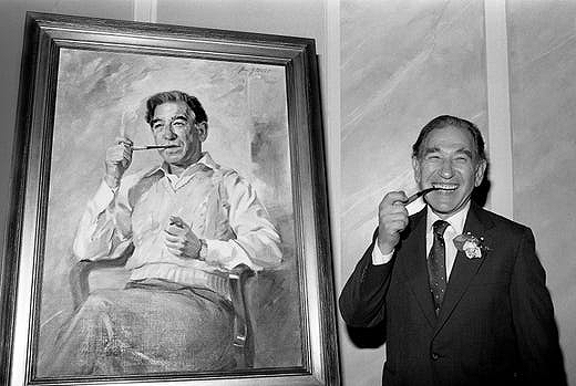 Stanley Cohen posing next to a portrait of himself. In the portrait, he is sitting down, smoking a pipe. His expression is contemplative and he is wearing slacks and a light-colored sweater vest over a white, collared shirt. Standing next to the painting, Cohen is holding his pipe to his mouth and is grinning at the camera. He is wearing a black suit with a white shirt and tie and has a flower pinned to his lapel. The photo is in black and white.