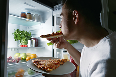 Young person at an open fridge at night. They are eating a slice of pizza off a plate with another piece on it.