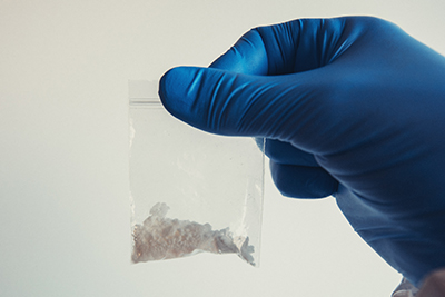 A gloved hand holding a little resealable baggie with cocaine inside it. The glove is blue and the background is white.