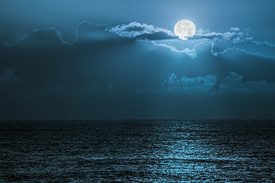A nighttime shot of the ocean, with the moon peeking out from behind some clouds. It is shining strongly and illuminates the water.