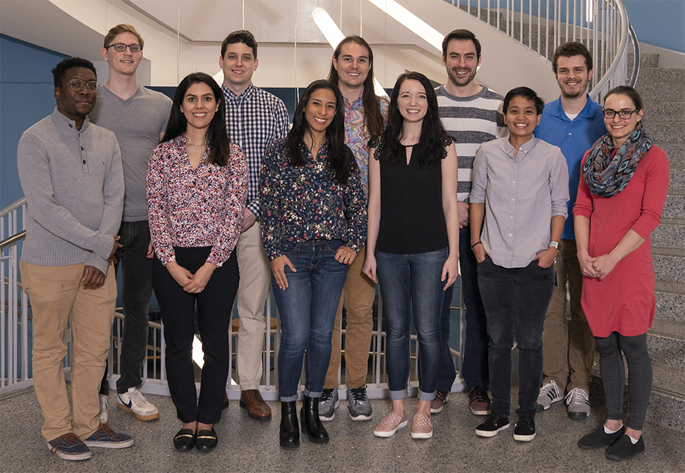 All eleven winners of the 2019 award, standing in a line, in the same order in which they are listed in the body of this webpage. They are standing in the stairwell of the Science and Engineering Building, with its white tube lights hanging above their heads.