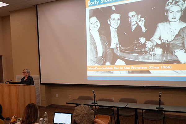 Linda Hughes speaking behind a prodium with a large photo of lesbian women drinking in a gay bar is on the screen