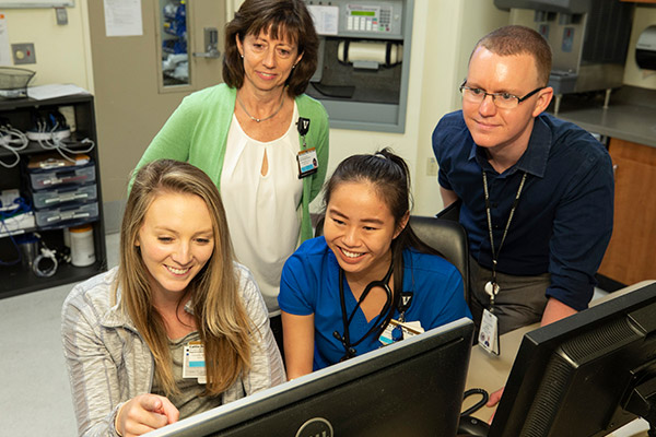 Two nurses seated at a computer monitor in a hospital nursing station while two informatics professionals, one female, one male, review the content on the monitor