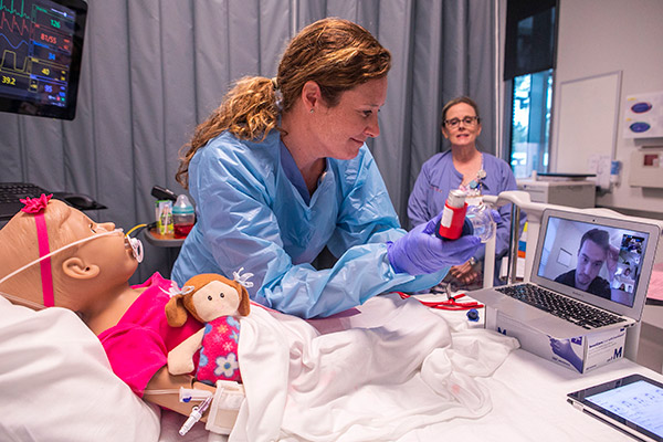 Vanderbilt nursing instructors in blue gowns communicate via laptop camera with a student about activity in VUSN's Simulation Lab