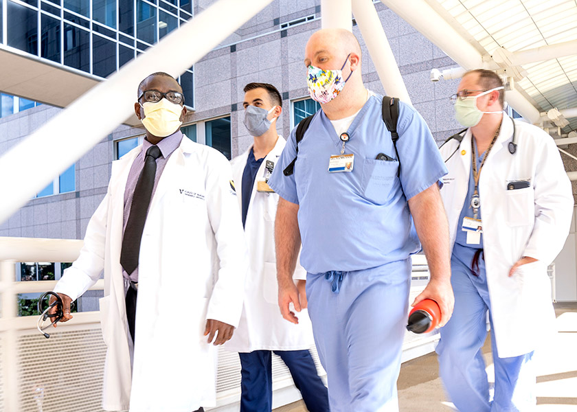 Four VUSN male students walk across a hospital skybridge; all wear masks and scrubs or whitecoats.